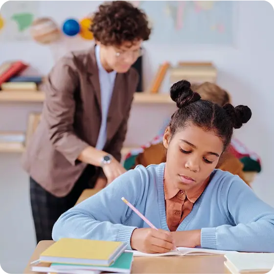 A focused middle school student writes in a notebook while a teacher supports another student in the background.